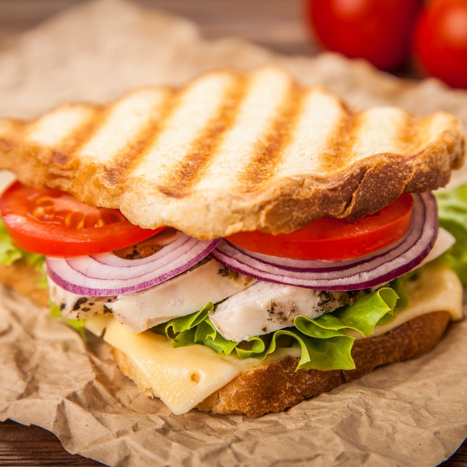 Grilled sandwich with chicken, cheese, lettuce, red onion, and tomato on crumpled brown paper.