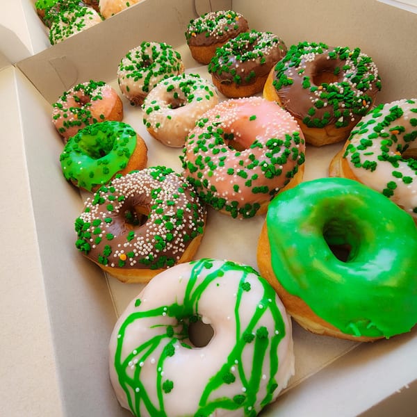 A box of KB Donuts with green, pink, and chocolate frosting, topped with green and white sprinkles.