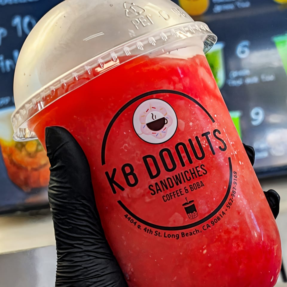 Hand in black glove holding a red KB Donuts drink with a clear domed lid.