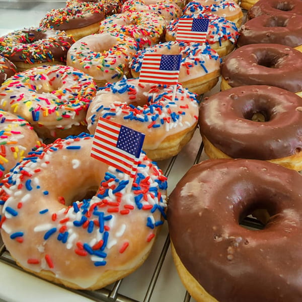 Rows of glazed and chocolate donuts, some with patriotic sprinkles and American flag picks.