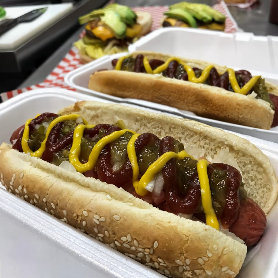 Two hot dogs with ketchup, mustard, relish, and onions in white foam containers, with burgers in the background.