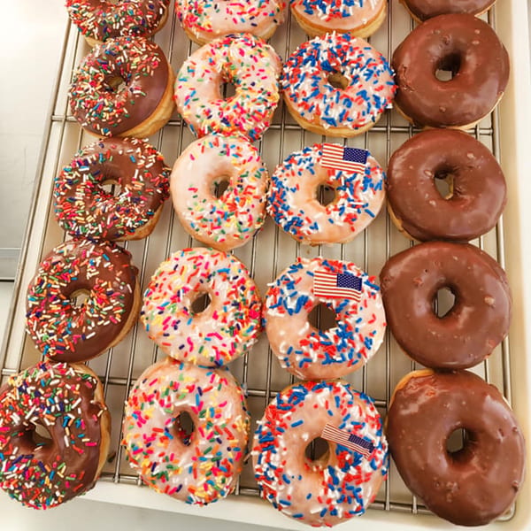 Assortment of KB Donuts, some with chocolate frosting and sprinkles, others with patriotic red, white, and blue sprinkles.