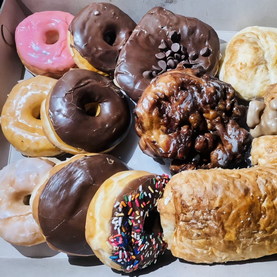 A box filled with various KB Donuts, including glazed, chocolate, pink, and sprinkle-covered donuts.